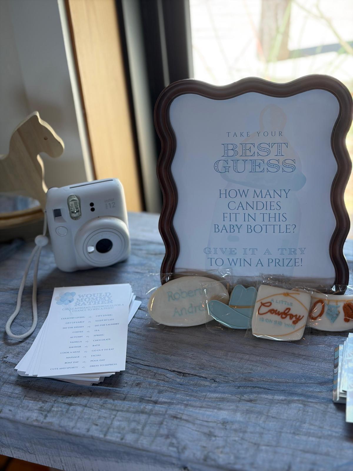 Framed sign inviting guests to guess how many candies fit in a baby bottle, beside decorated cookies
